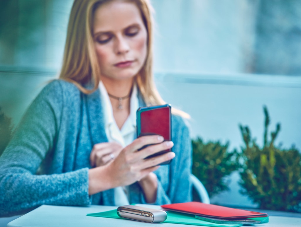 Blond woman checking her phone with a IQOS device on the table