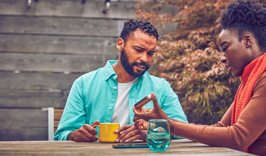 Two people at a table bar, one holds his IQOS device and the other uses his phone