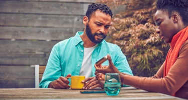 Two people at a table bar, one holds his IQOS device and the other uses his phone